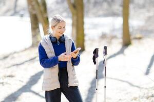 Young hiker woman using smartphone and listening music with headphones and trekking poles in forest photo