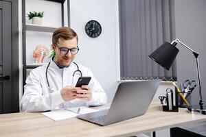 Male doctor medical worker in modern clinic wearing eyeglasses and white coat uniform using cell mobile smartphone apps, sitting at laptop computer photo