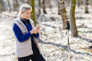 Young hiker woman using smartphone and listening music with headphones and trekking poles in forest. photo