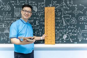 Asian male math teacher holding book and large ruler standing in front of chalkboard with formulas and geometry photo