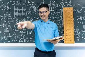Asian math teacher smiling and pointing forward holding book in classroom with blackboard of formulas and geometry photo