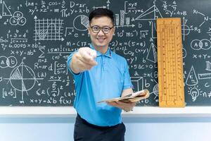 Asian math teacher pointing chalk at camera holding book standing in front of blackboard with formulas and geometric drawings photo