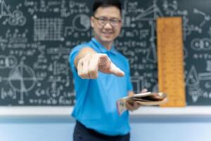 Asian math teacher pointing directly at camera holding book standing in classroom with blackboard of formulas and geometry photo