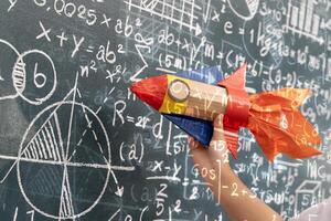 Hand holding colorful rocket craft in front of blackboard full of math and science formulas with overlay effect photo