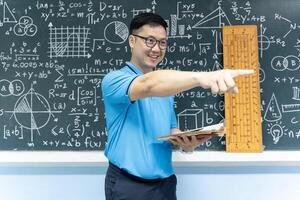 Asian math teacher smiling and pointing to side holding book standing in classroom with blackboard of formulas and drawings photo