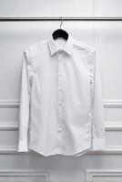 A crisply ironed, pristine white collared shirt hangs neatly on a black hanger against a textured white wall. photo