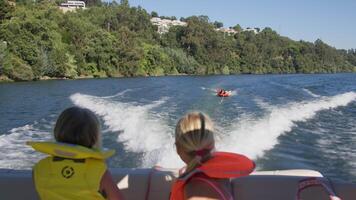 Children wearing life vests sitting in a boat watching a person riding an inflatable tube on a river surrounded by nature video