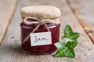 Jar of homemade jam with a rustic burlap lid and a handwritten label, placed on a wooden surface alongside fresh green ivy leaves, showcasing natural food presentation photo