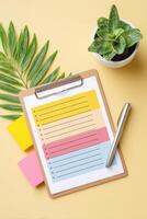 Colorful clipboard with checklist paper, sticky notes in vibrant colors, and a small potted plant on a yellow background, creating an organized workspace atmosphere photo