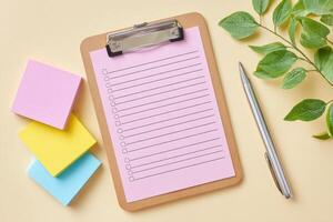 Clipboard with pink checklist paper, colorful sticky notes in pastel shades, silver pen, and green leaves on a soft beige background, creating an organized workspace atmosphere photo