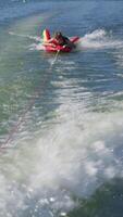 Teenager tubing on a colorful inflatable towed by a boat, creating water splashes and wake trails on the lake surface video