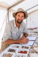 Male geologist wearing a hat is studying mineral samples on a table in a tent, with various specimens displayed, showcasing the process of geological research and analysis photo