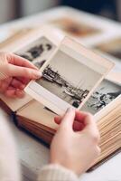 Individual holding a vintage photograph of boats on water, surrounded by an open photo album filled with black and white images, evoking nostalgia and memories of the past