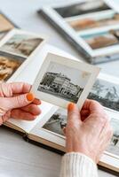 Hands holding a vintage black and white photograph of a historical building, surrounded by an open photo album filled with various images, evoking nostalgia and memories