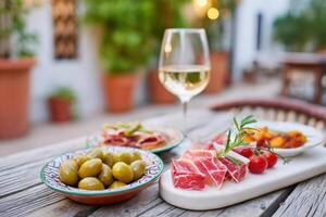 Elegant table setting featuring assorted appetizers including olives, cured meats, and fresh tomatoes, accompanied by a glass of white wine in a cozy outdoor dining atmosphere photo