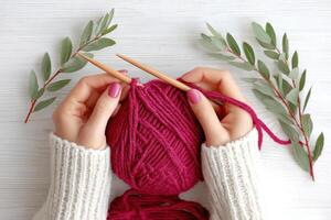 Hands of a woman holding knitting needles, working with a vibrant pink yarn ball surrounded by green leaves, showcasing a cozy crafting atmosphere and creative hobby photo