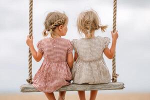 Two young girls sitting on a wooden swing, wearing matching dresses, enjoying a serene moment outdoors, with a soft cloudy sky in the background, evoking childhood innocence and joy photo