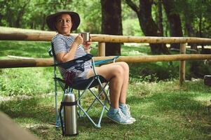 Woman Relaxing Outdoors in a Camping Chair Holding a Hot Drink in a Serene Forest Setting photo