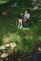 Woman Relaxing Outdoors Using a Laptop in a Natural Setting by a Calm Stream photo