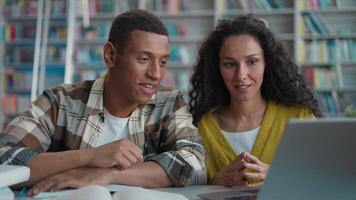 Two students African American man Hispanic Arabian woman together project teamwork discuss talking classmates laptop computer library university studying cooperation female male learning high school video