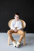 An 11 year old boy with reading glasses and hearing aids in a white shirt sits on a table against a black wall and smiles while looking at the frame photo