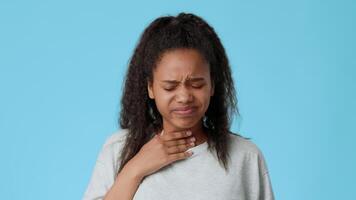 A young African American girl with curly hair appears to be feeling unwell. She stands against a bright blue backdrop and touches her throat, displaying a look of concern or discomfort. video