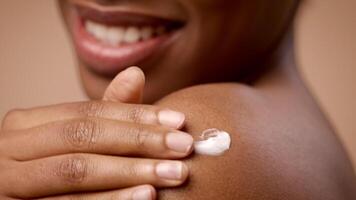 A pretty, short-haired African American woman is applying lotion to her shoulder. She is looking towards the camera with a smile, highlighting her skincare routine and natural beauty. video