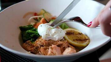 Toast with salmon, poached egg and avocado on white plate. The cook cuts the poached egg with a knife and fork. The yolk flows out. video