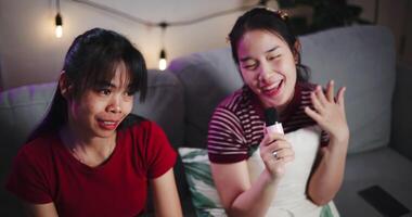 Handheld high angle view shot, Two Asian women sitting on a sofa happily singing karaoke with smiles and enjoying a fun time together at home. Share good time video
