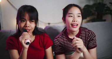 Handheld shot, Two Asian women sitting on a sofa happily singing karaoke with smiles and enjoying a fun time together at home. Enjoy a warm moment video