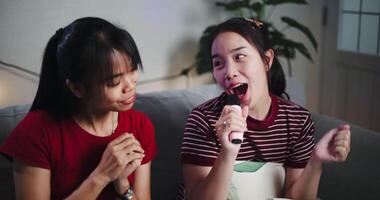 Handheld shot, Two Asian women sitting on a sofa happily singing karaoke with smiles and enjoying a fun time together at home. Share good time video