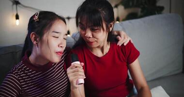 Handheld high angle view shot, Two Asian women sitting on a sofa happily singing karaoke with smiles and enjoying a fun time together at home. video