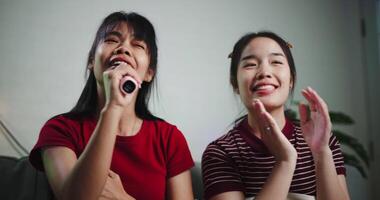 Handheld low angle view shot, Two happy women sitting on a sofa happily singing karaoke with smiles and enjoying a fun time together at home. Share good times video