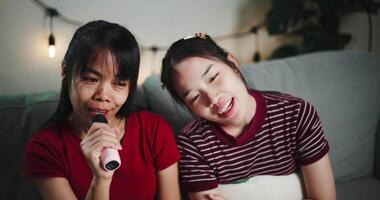 Handheld high angle view shot, Two Asian women sitting on a sofa happily singing karaoke with smiles and enjoying a fun time together at home. Enjoy a warm moment video