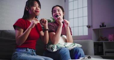 Handheld shot, Two Asian women sitting on a sofa happily singing karaoke with smiles and enjoying a fun time together at home. video