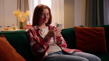 Positive happy smiling laughing Caucasian woman female girl user gadget addict using online app smartphone social media chat communication sitting on home couch typing message mobile phone cheerful video