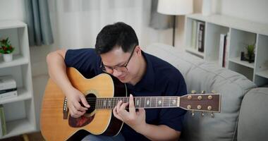 Handheld high angle view shot, Relaxed man wearing glasses and a navy-blue shirt is enjoy playing an acoustic guitar on a sofa in the living room at home. video