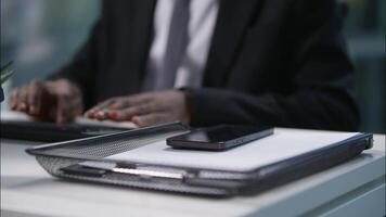 Close up workplace documents desktop African American man holding chatting mobile phone smartphone multitasking using computer online businessman male guy business office worker employer entrepreneur video