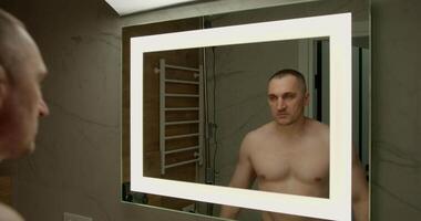 man stands shirtless in front of a mirror flexing his muscles and gazing intently at his reflection. The modern bathroom features soft lighting and clean lines enhancing the focused atmosphere video