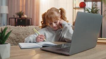 Tired young girl doing homework at table with laptop writing in notebook and showing lazy expression video