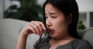 Young Asian woman sit on the sofa alone, lost in thought. Her brows were slightly furrowed and expression tense in the quiet atmosphere at home video