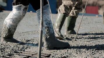 Close-up of workers in rubber boots standing in wet concrete during slab pouring, Protective wrapping on legs for safety during manual construction process video