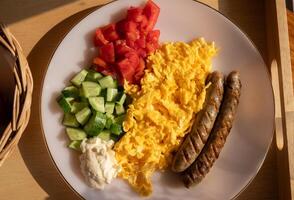 Scrambled eggs with sausage and pieces of tomato and cucumber served on a dish as breakfast. Breakfast is the first meal of the day usually eaten in the morning. photo