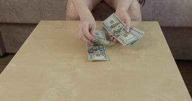 Female hands are seen counting stack of US dollar bills on wooden table setting is cozy indoor space suggesting personal or casual environment video