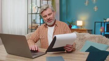Mature man sitting on table with documents and laptop, smiling from successful work at home office video