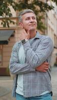Thoughtful smiling mature man with pensive expression touching chin looking around on city street video