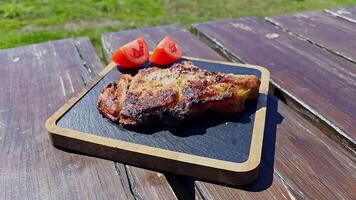 Grilled pork steak served with fresh tomato wedges on wooden tray, juicy barbecue meat dish presented outdoors on rustic table under sunny weather video