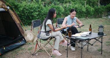 Handheld high angle view shot, Two Asian women sit together in a garden, happily chatting while enjoying snacks and drinks, surrounded by a cozy camping vibe. Holiday Activities video