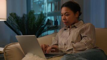 Focused African American woman ethnic girl 30s female working with laptop on comfortable couch use computer device on sofa browsing internet social media studying shopping online in night evening home video