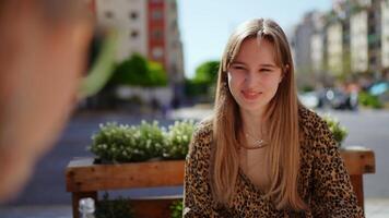 Cheerful young woman sits at an outdoor cafe, engaging in a pleasant conversation with someone off screen, her expressive face reflecting enjoyment and connection video